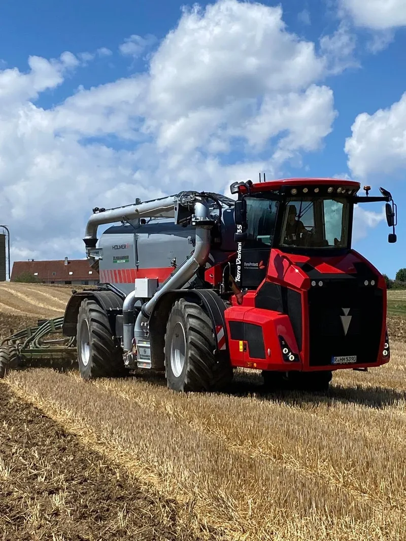 Roter Gülleselbstfahrer mit Aufbaufass auf einem Stoppelfeld unter blauem Himmel